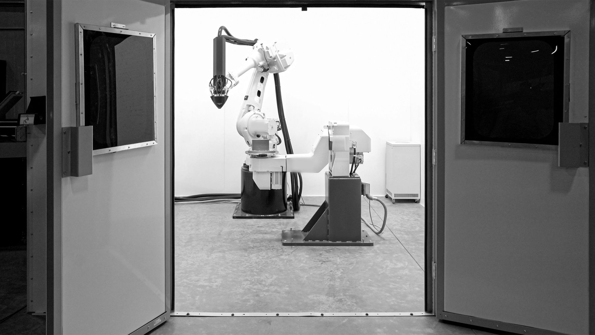 A robotic arm in a white work cell, seen through a pair of open doors